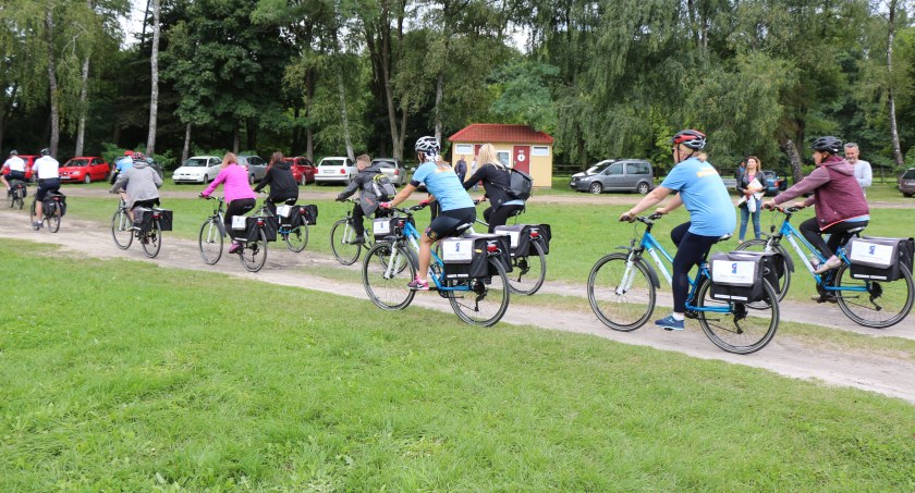 Zdjęcie: Grupa rowerzystów obojga płci jedzie nieutwardzoną drogą. W tle widać parking z kilkoma samochodami, a za nim liściasty las